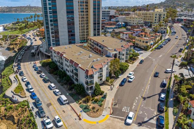 An aerial view of a coastal city street lined with multi-story residential apartment buildings and parked cars. On the left, a road runs parallel to a rocky ocean coastline with crashing waves, while a taller high-rise building stands prominently in the background.
